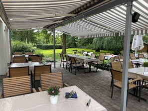Un patio exterior con mesas y sillas, cubierto por toldos a rayas, rodeado de vegetación.