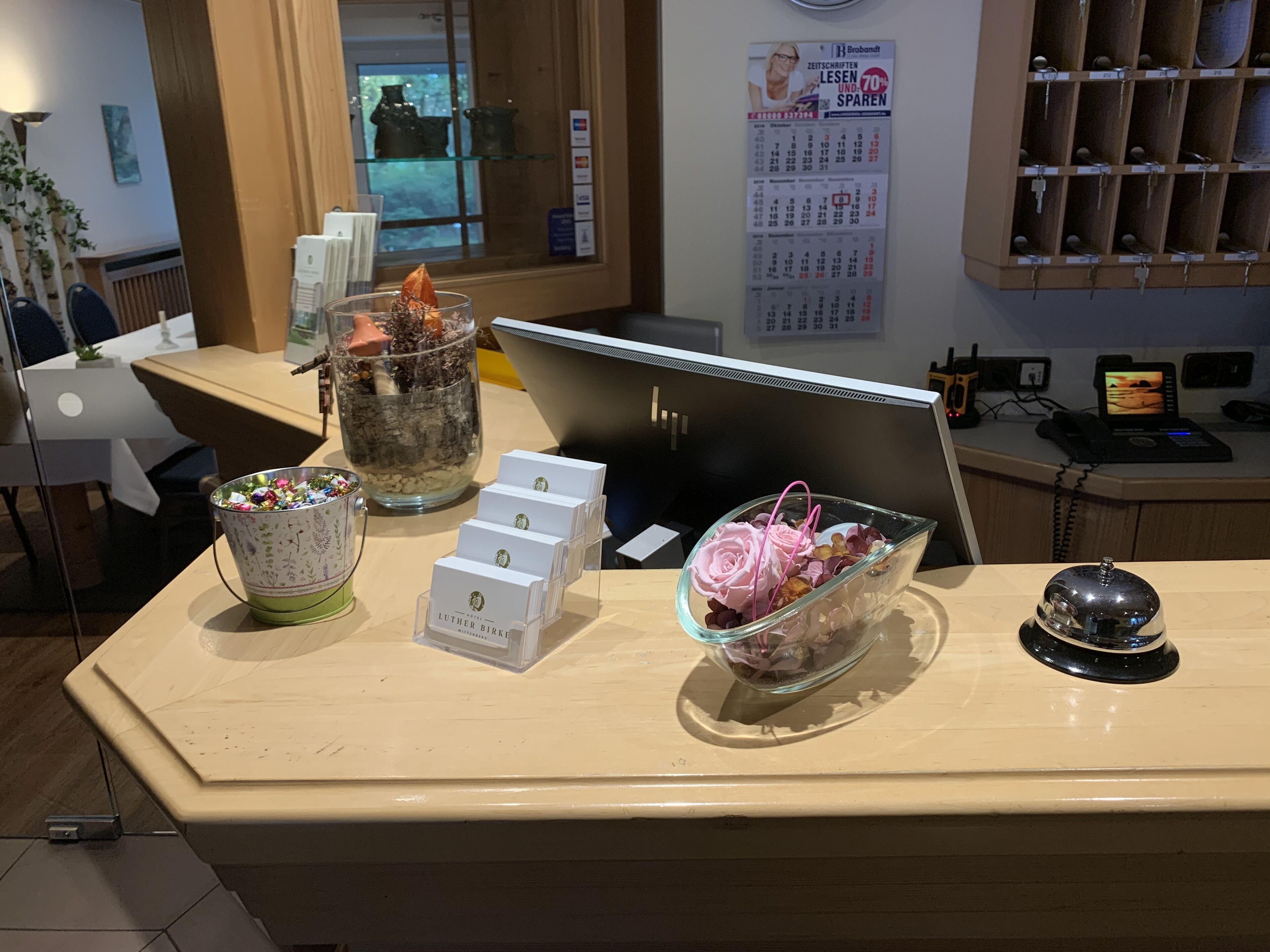 Un mostrador de recepción con un calendario, un florero de vidrio con flores, una taza de café y una planta pequeña.
