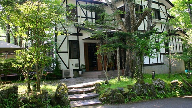 A traditional half-timbered house with a stone staircase leading to the entrance, surrounded by lush greenery and trees. - Pension Marie (Yamanakako)