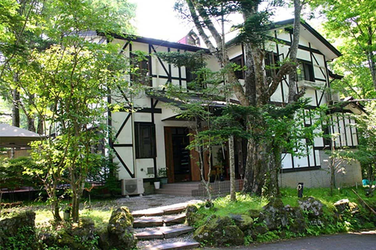 A traditional half-timbered house with a stone staircase leading to the entrance, surrounded by lush greenery and trees.