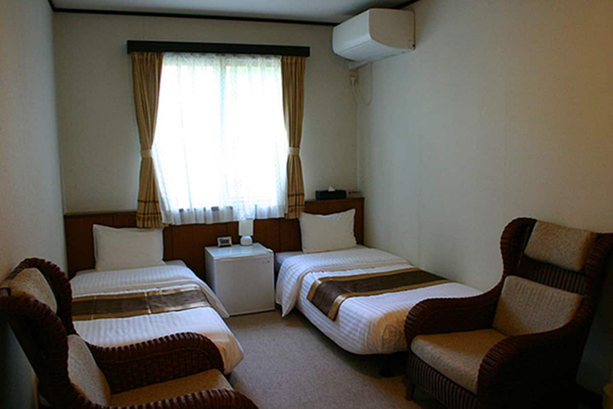 A hotel room with two beds, a small table, a chair, and an air conditioning unit.