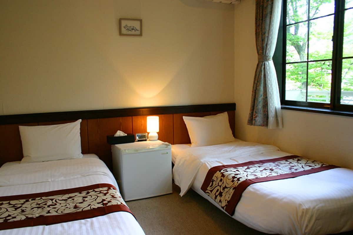 A hotel room with two beds, a bedside table with a lamp, a small refrigerator, and a window with curtains.
