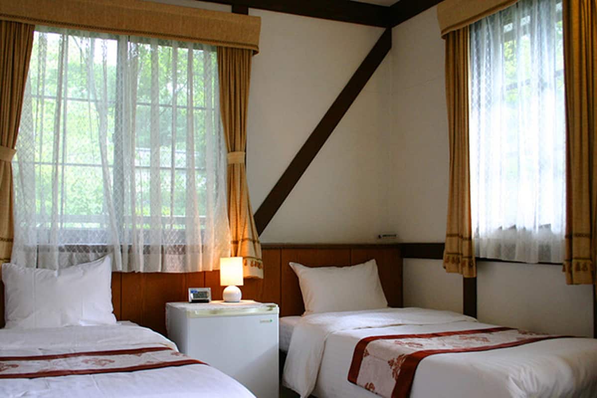 A room with two beds, a nightstand with a lamp, and windows with curtains.