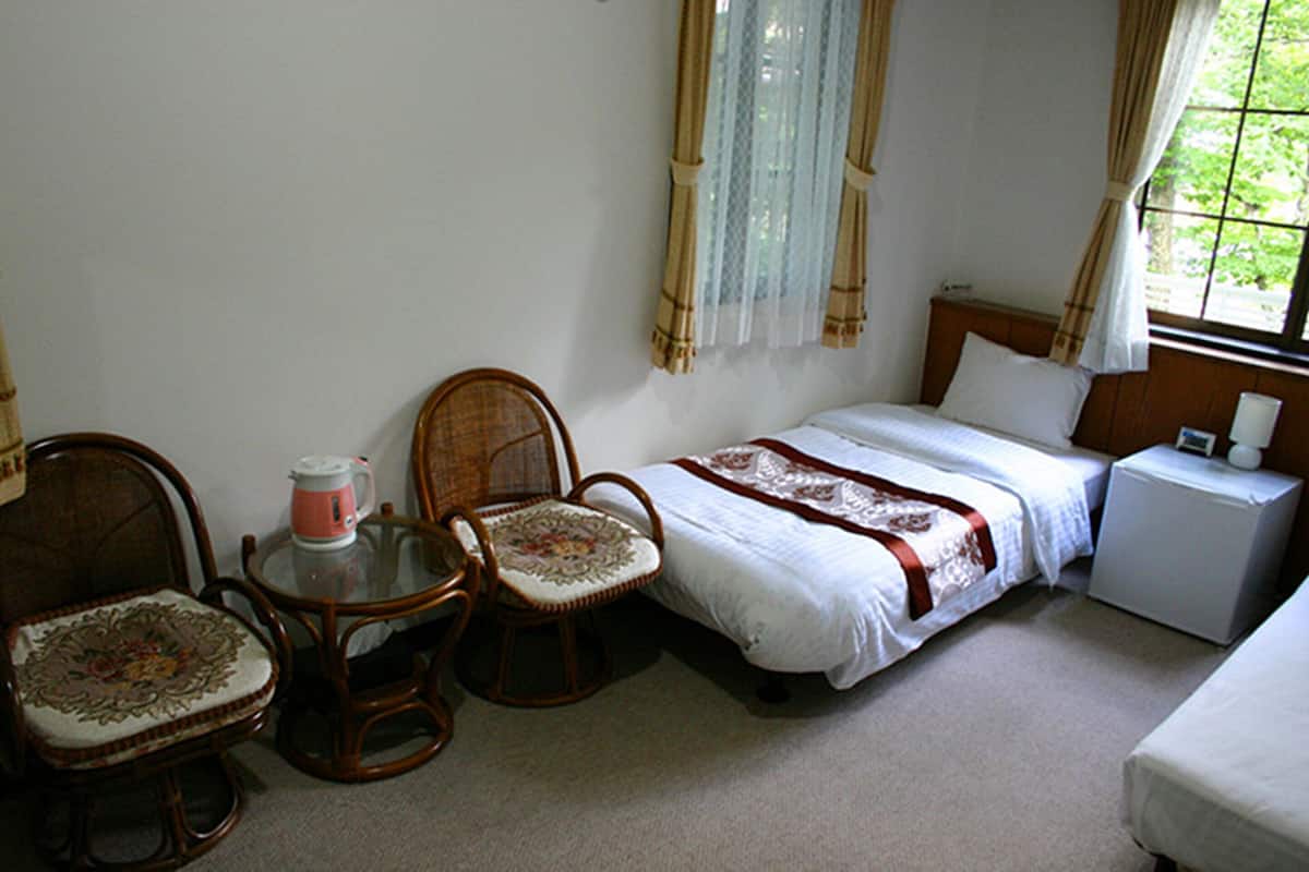 A bedroom with a bed, two chairs, a small table with a kettle, and a nightstand with a lamp.