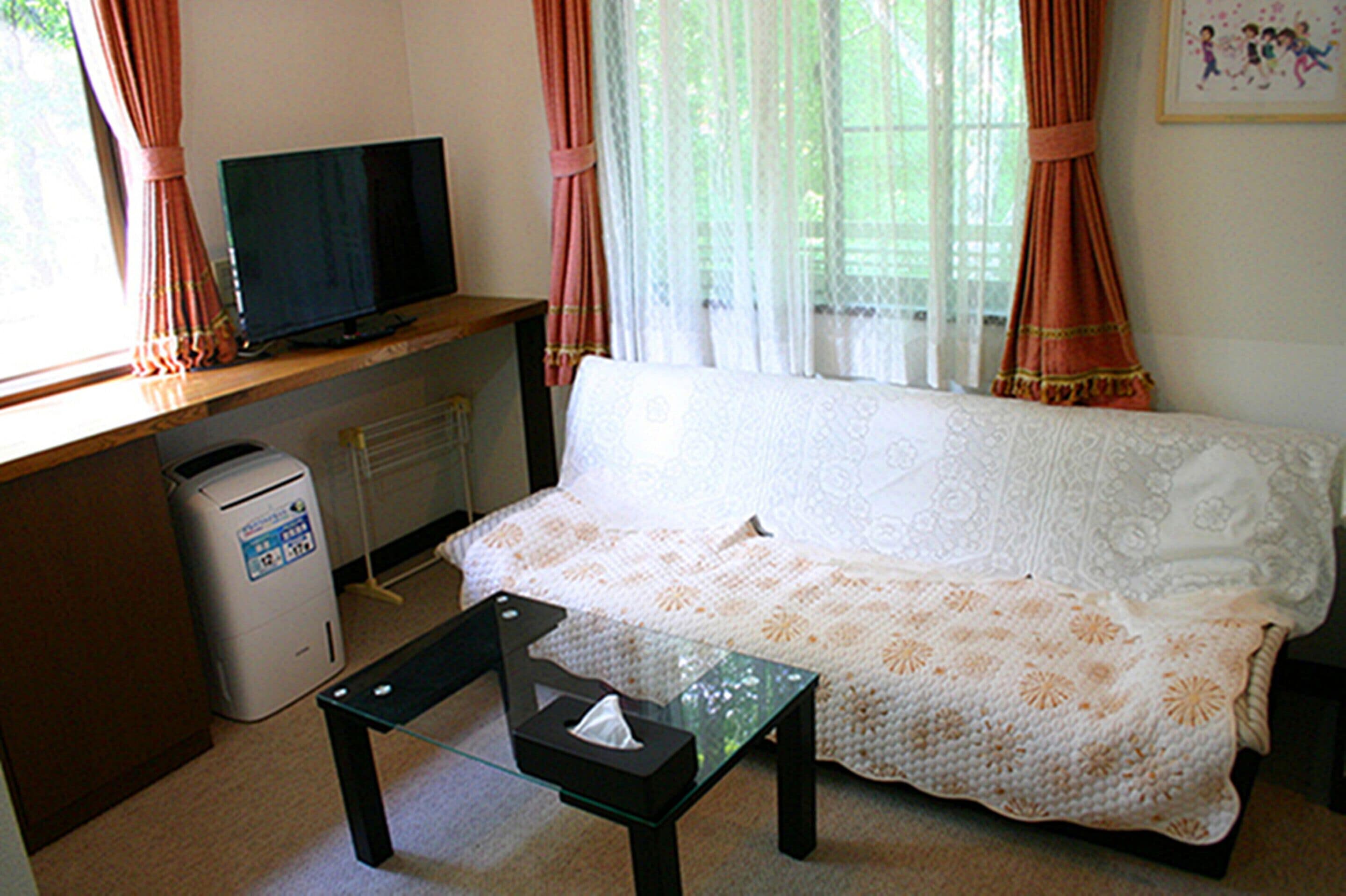A living room with a sofa, a glass coffee table, a television on a wooden stand, a waste bin, and a window with curtains.