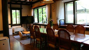 A dining area with a wooden table and chairs, a television, and large windows with curtains. - Pension Marie (Yamanakako)