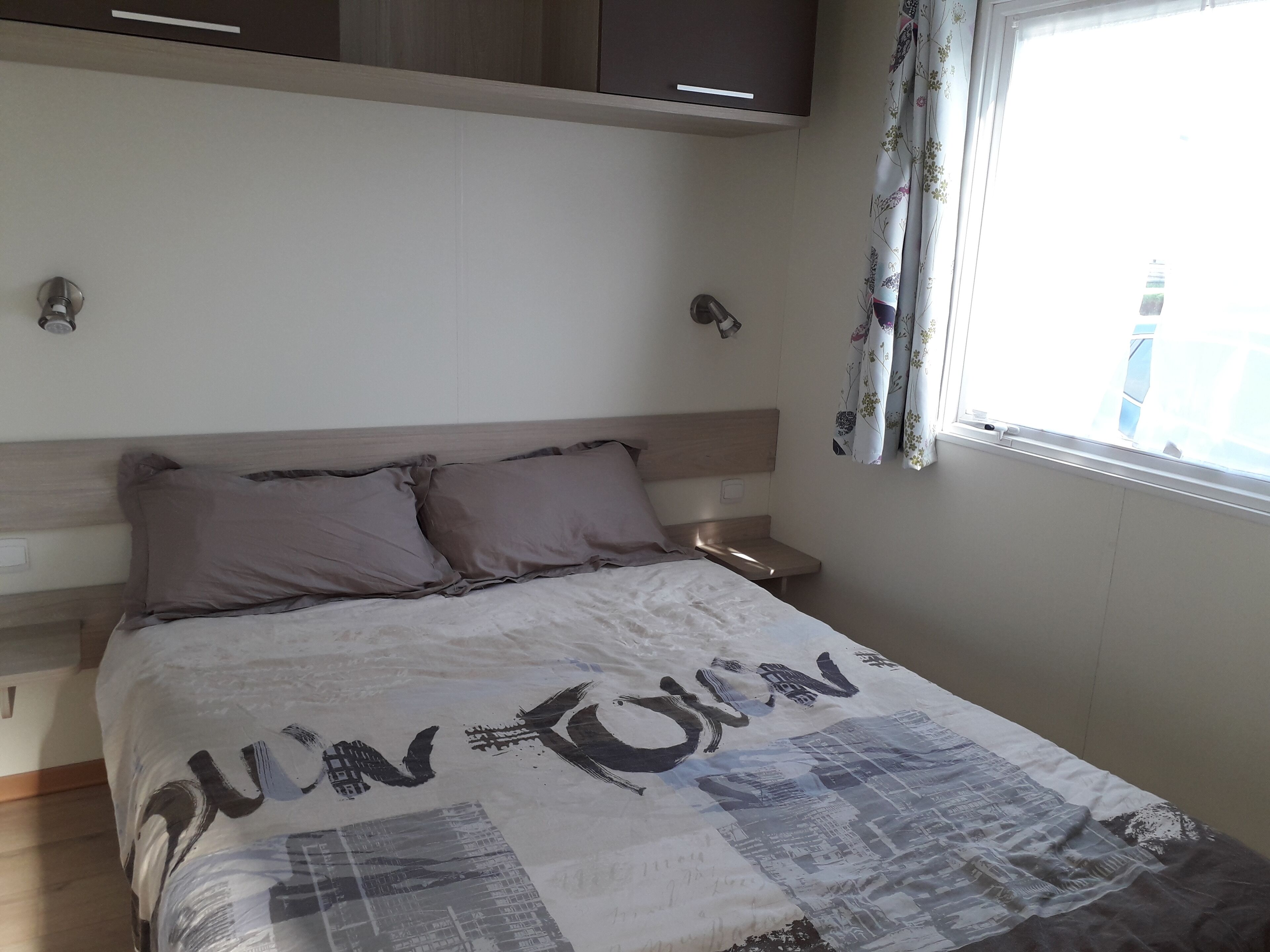 Une chambre à coucher avec un lit, une table de chevet, une fenêtre avec des rideaux et une tête de lit en bois.