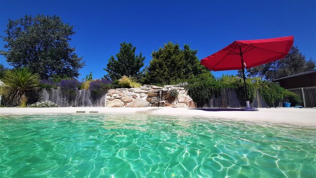 A clear pool with a stone wall and waterfall feature, surrounded by lush greenery and a red umbrella.
