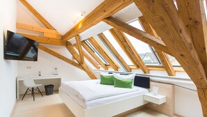 A modern bedroom with a sloped ceiling, wooden beams, a bed with white bedding, a desk with a chair, and a TV mounted on the wall. - DIE KRONE - Hotel Garni (Kirchzarten)