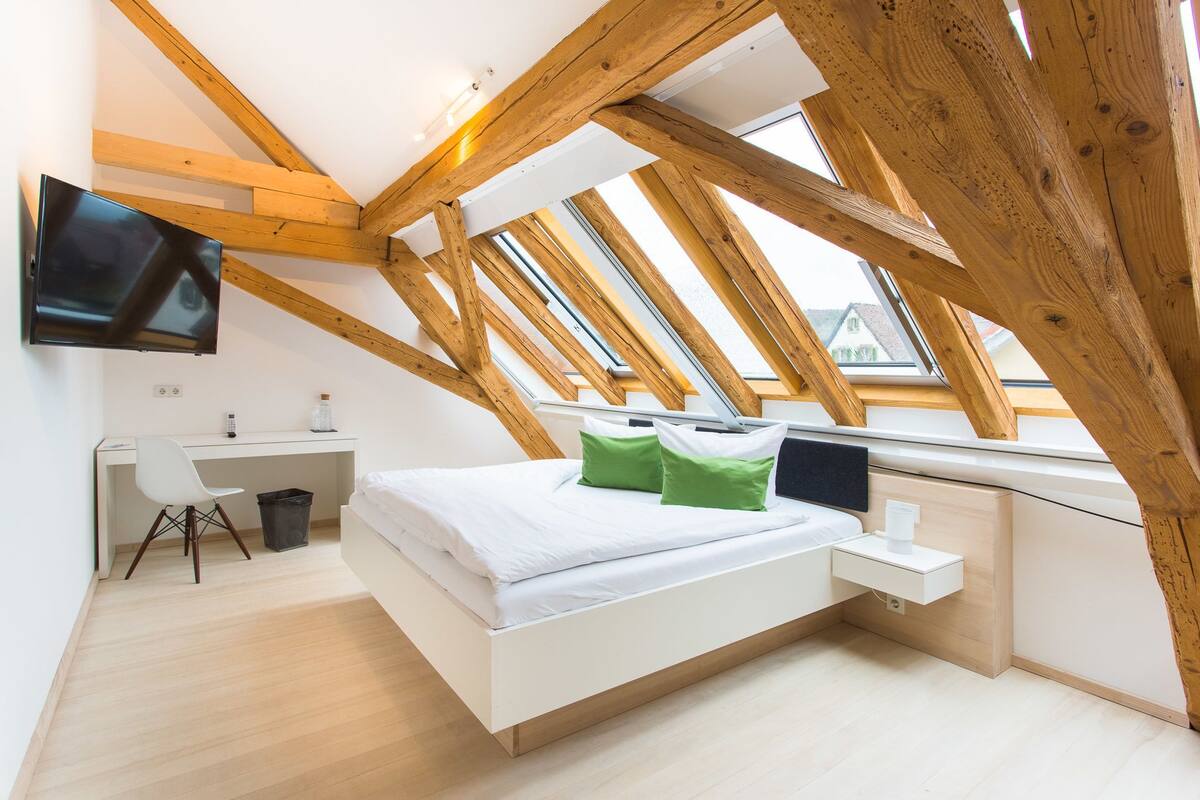 Een moderne slaapkamer met een schuine kap, houten balken, een bed met witte beddengoed, een bureau met een stoel en een tv aan de muur.