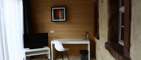 Una habitación pequeña con un escritorio blanco, una silla blanca, un televisor sobre un mueble bajo, un cuadro en la pared y una ventana con vistas al exterior.