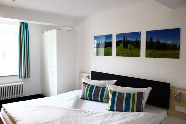 Habitación de hotel con cama, mesita de noche, radiador y tres cuadros de paisajes en la pared.