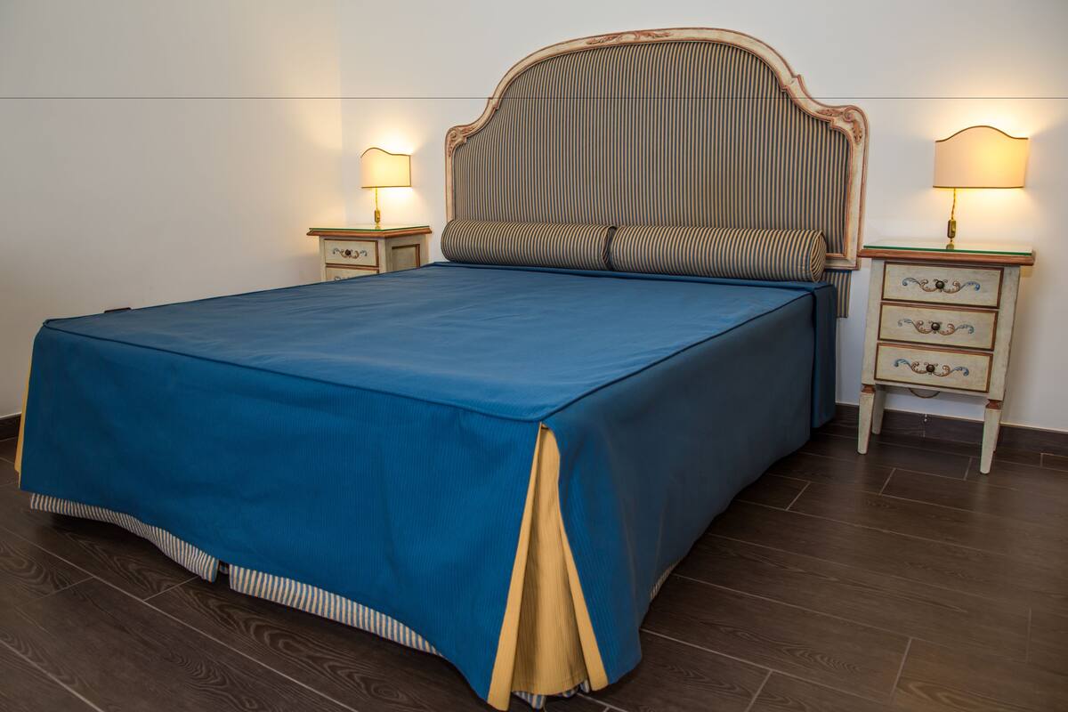 A neatly made bed with a blue bedspread and a wooden headboard. There are two bedside tables with lamps on either side of the bed.