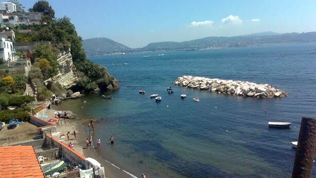 A coastal town with a rocky breakwater, clear blue sea, and buildings on a hillside. - La Rada B&B (Bacoli)