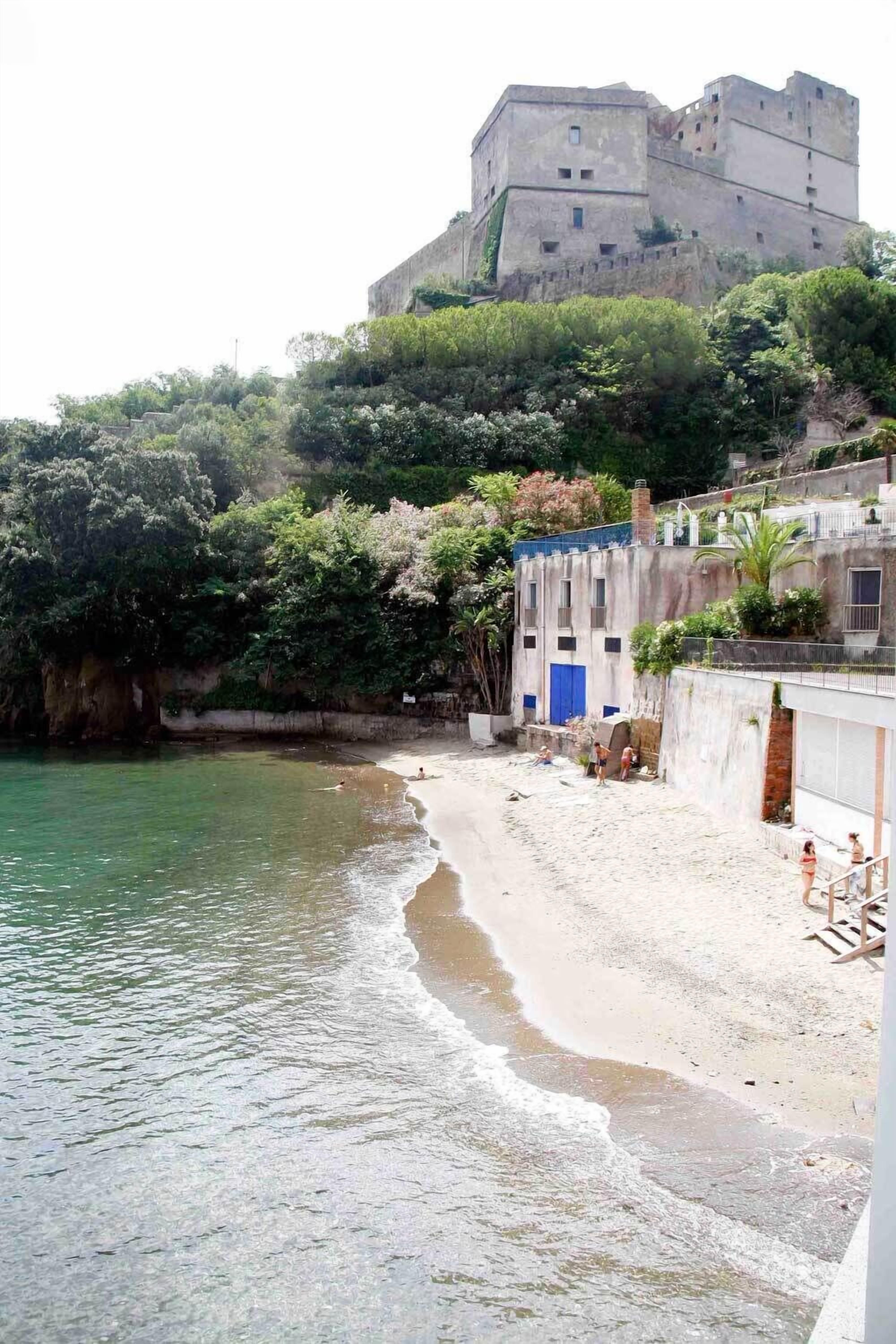 Uma paisagem costeira com uma praia de areia, um edifício de pedra com porta azul e uma construção semelhante a um castelo numa colina.