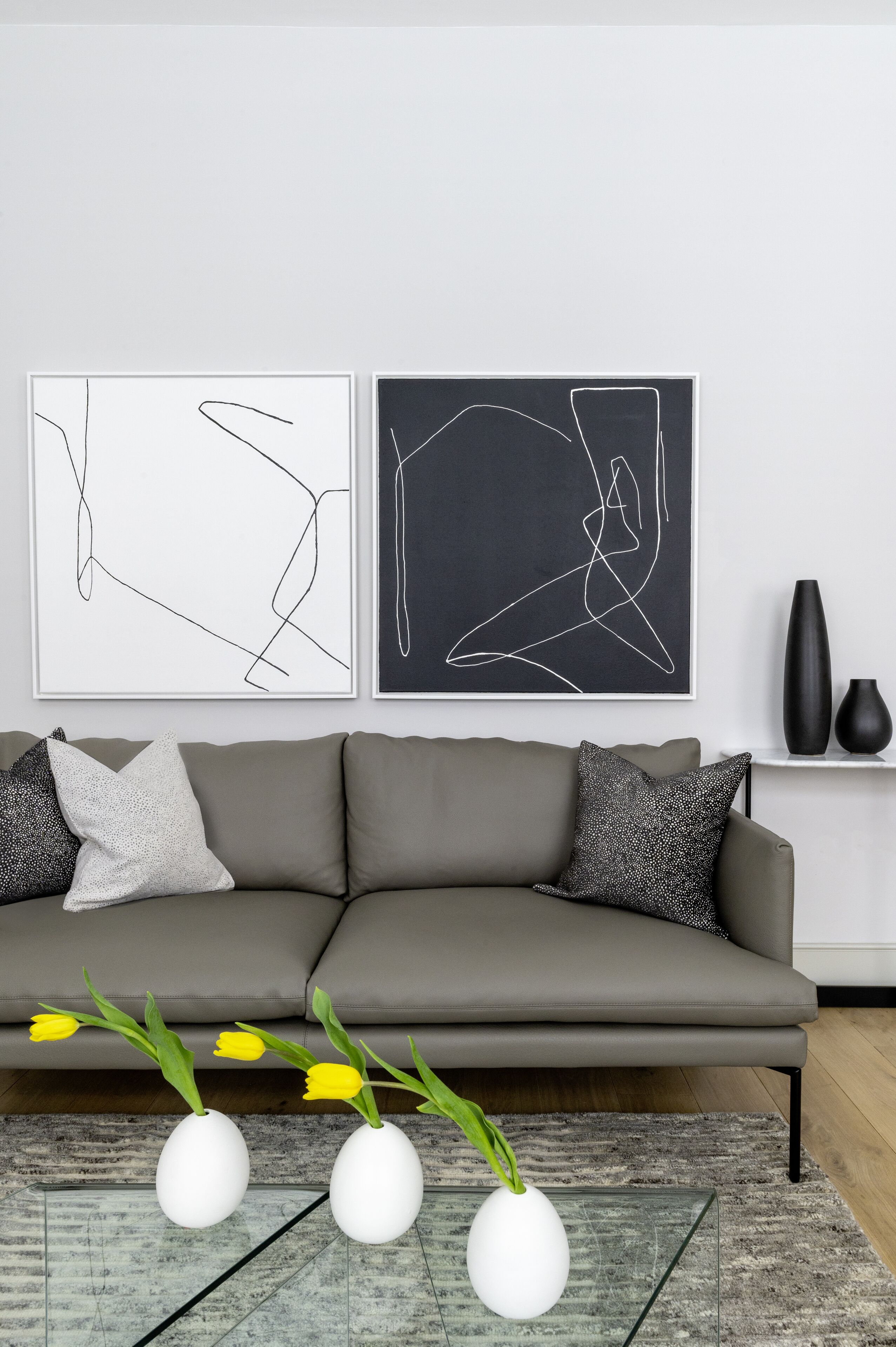 A modern living room with a grey sofa, two abstract wall art pieces, and three white vases with yellow flowers on a glass table.