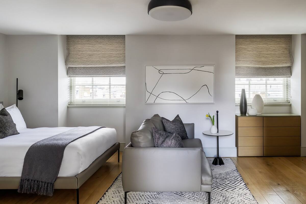 A modern bedroom with a bed, a sofa, a small side table, and a window with blinds.