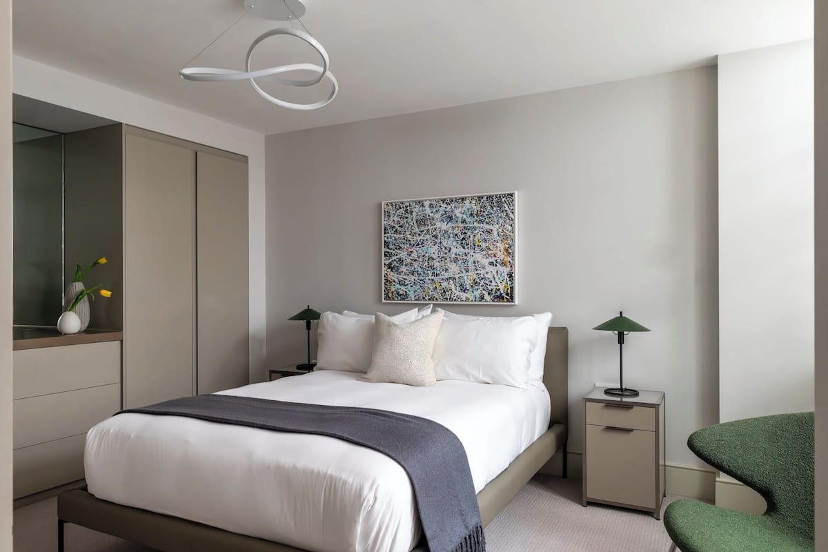 A modern bedroom with a large bed, a nightstand, a green chair, and a wall-mounted abstract painting.