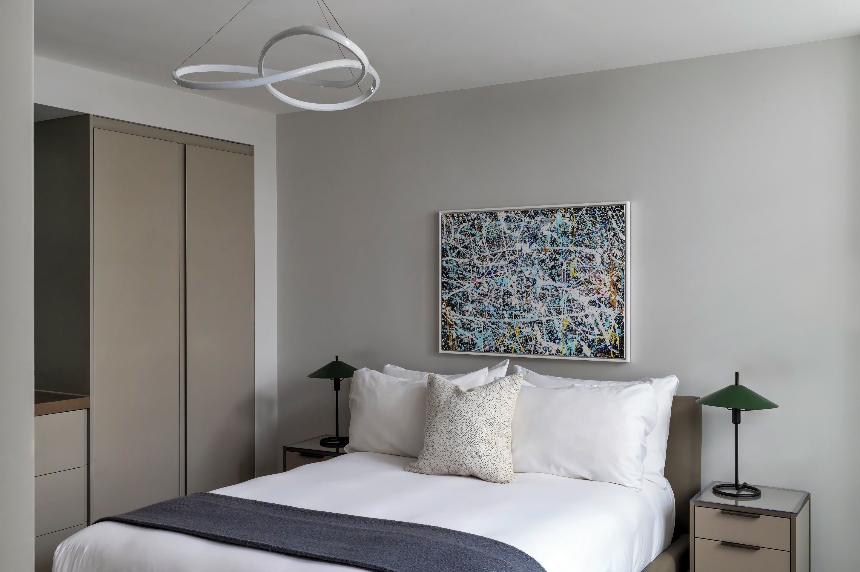 Une chambre moderne comprenant un lit, deux tables de chevet, une commode, une armoire, un luminaire au plafond et une œuvre d’art murale.