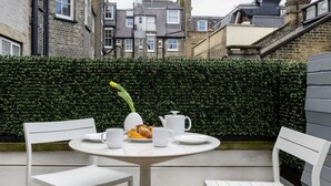 A round table with white chairs, a vase with a flower, and a croissant on a wooden deck. - AKA Marylebone (London)