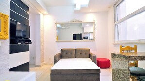 Una sala moderna con sofá cama, televisor, mesa de comedor y una cocina visible a través de una puerta abierta.
