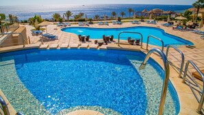 A swimming pool with lounge chairs and umbrellas, surrounded by palm trees and a view of the sea.