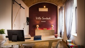 A room with a desk, chairs, a laptop, a TV, a small table with a candle, and a wall with a sign reading "Villa Santadi". - Villa Santadi Hotel (Santadi)