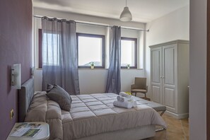 A bedroom with a bed, a chair, a wardrobe, and two windows with curtains. - Villa Santadi Hotel (Santadi)