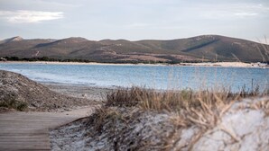 A coastal landscape with a wooden boardwalk leading to a sandy beach, surrounded by hills and a calm sea. - Villa Santadi Hotel (Santadi)