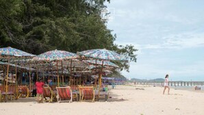 A beach with colorful umbrellas and beach chairs, a pier in the background, and a woman walking on the sand. - Rayong by Milanee (Rayong)