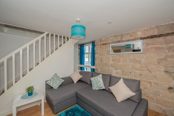 A modern living room with a grey sectional sofa, a white side table with a plant, a blue rug, a staircase with white railings, and a stone wall.