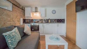 A compact living space with a kitchenette, a sofa with cushions, a small table with a glass, and a clock on the wall. - Kirkstall Bridge Apartments - 23 (Leeds)