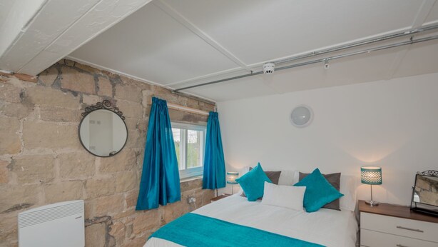 A bedroom with a stone wall, a bed with white and teal bedding, a round mirror, and a nightstand with a lamp. - Kirkstall Bridge Apartments - 23 (Leeds)