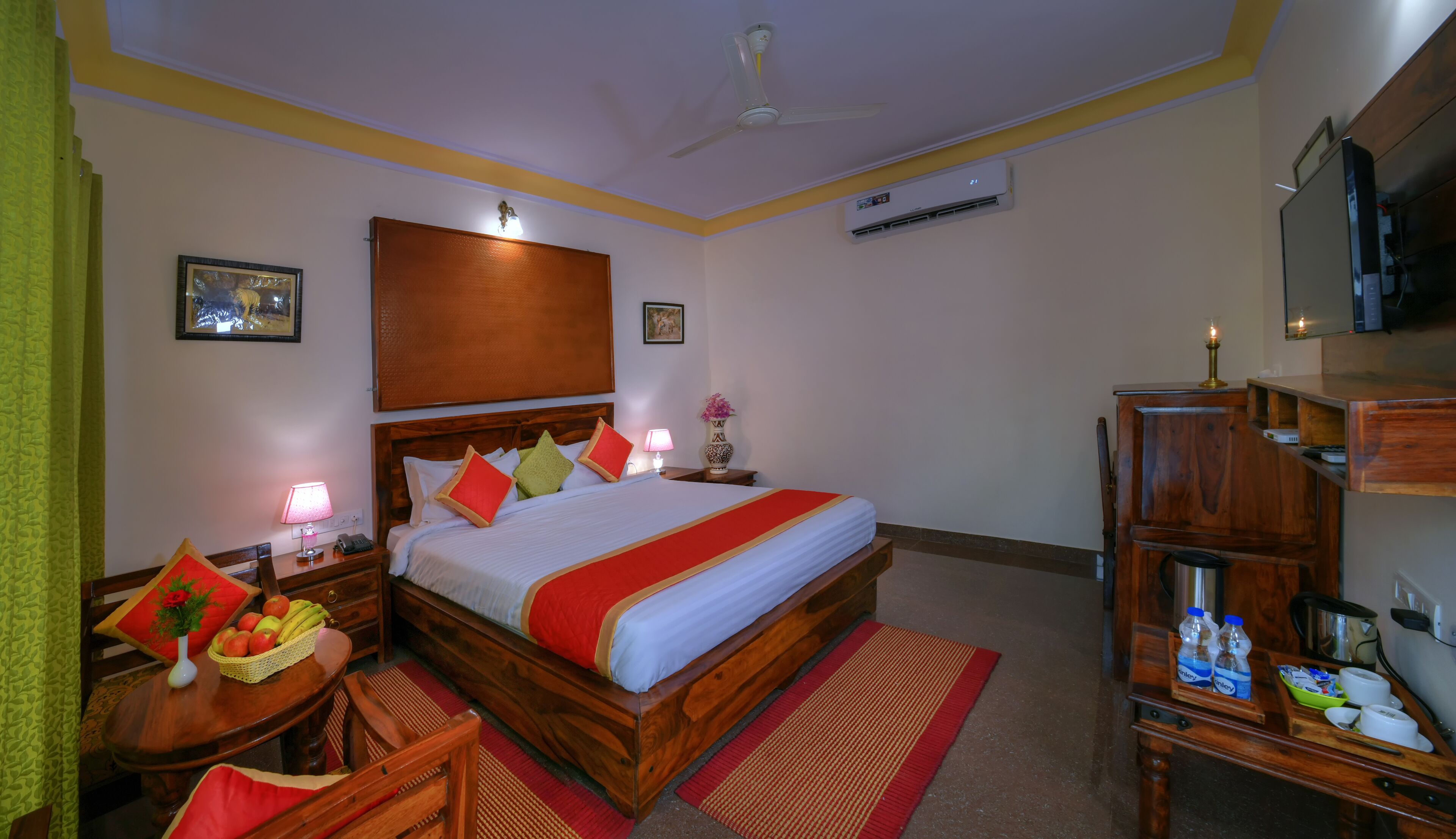 A hotel room with a wooden bed, a desk with a lamp, a chair, a small table with fruit, a TV, and an air conditioning unit.