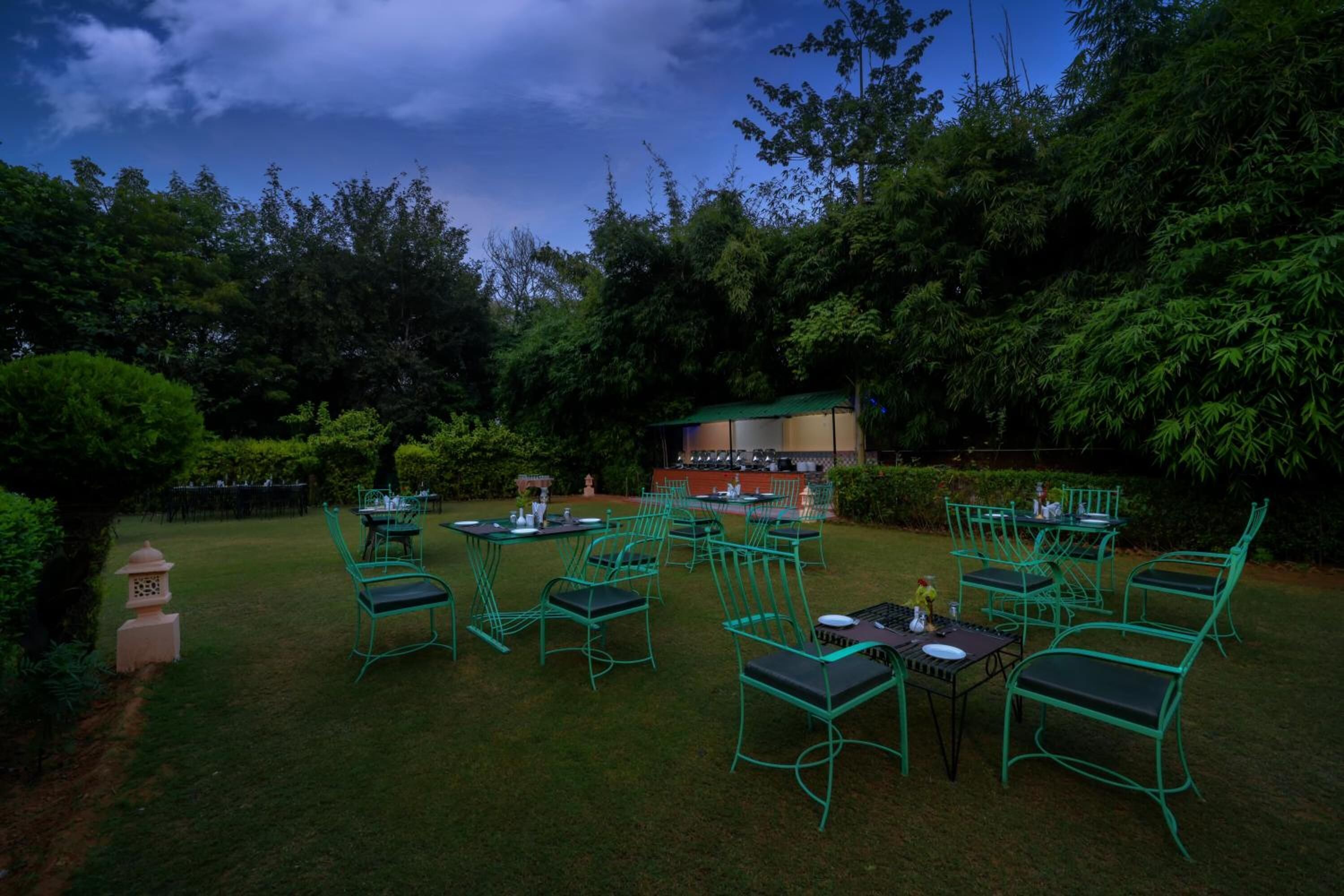 An outdoor dining area with green metal chairs and tables, surrounded by lush greenery and trees.