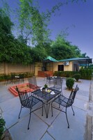 Un patio al aire libre con sillas y mesas de metal, una zona de barbacoa de ladrillo y un área de estar cubierta con techo verde.