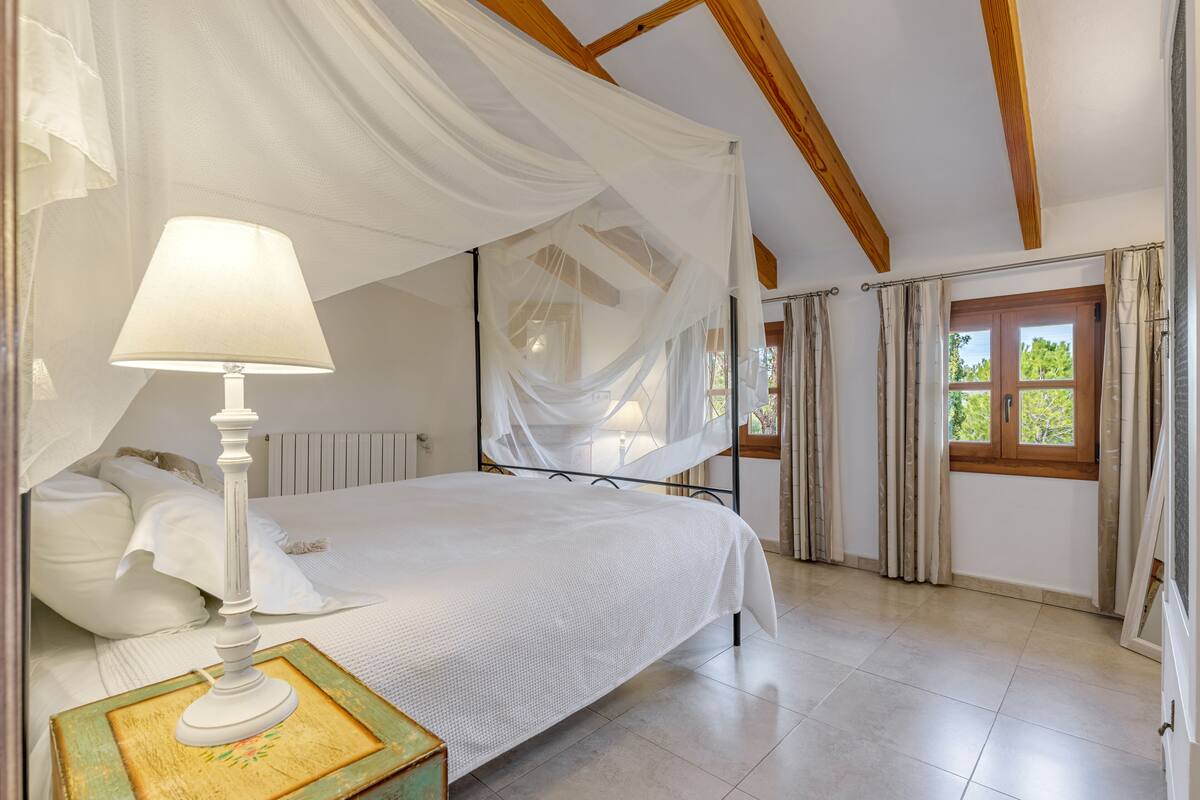 Quarto com cama de dossel, mesa de cabeceira com abajur e vista para o exterior pela janela.