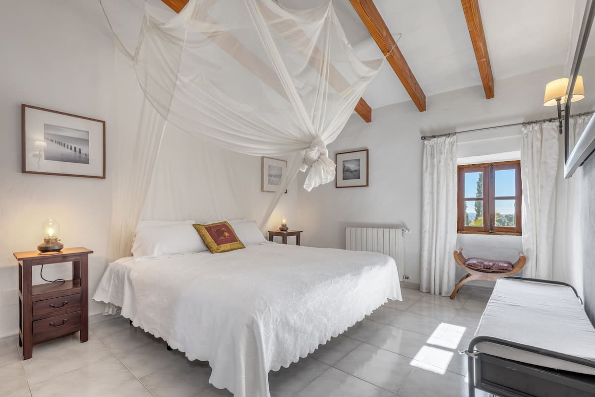 Um quarto com uma cama grande, uma mesa de cabeceira com abajur, uma janela com cortinas e um radiador.