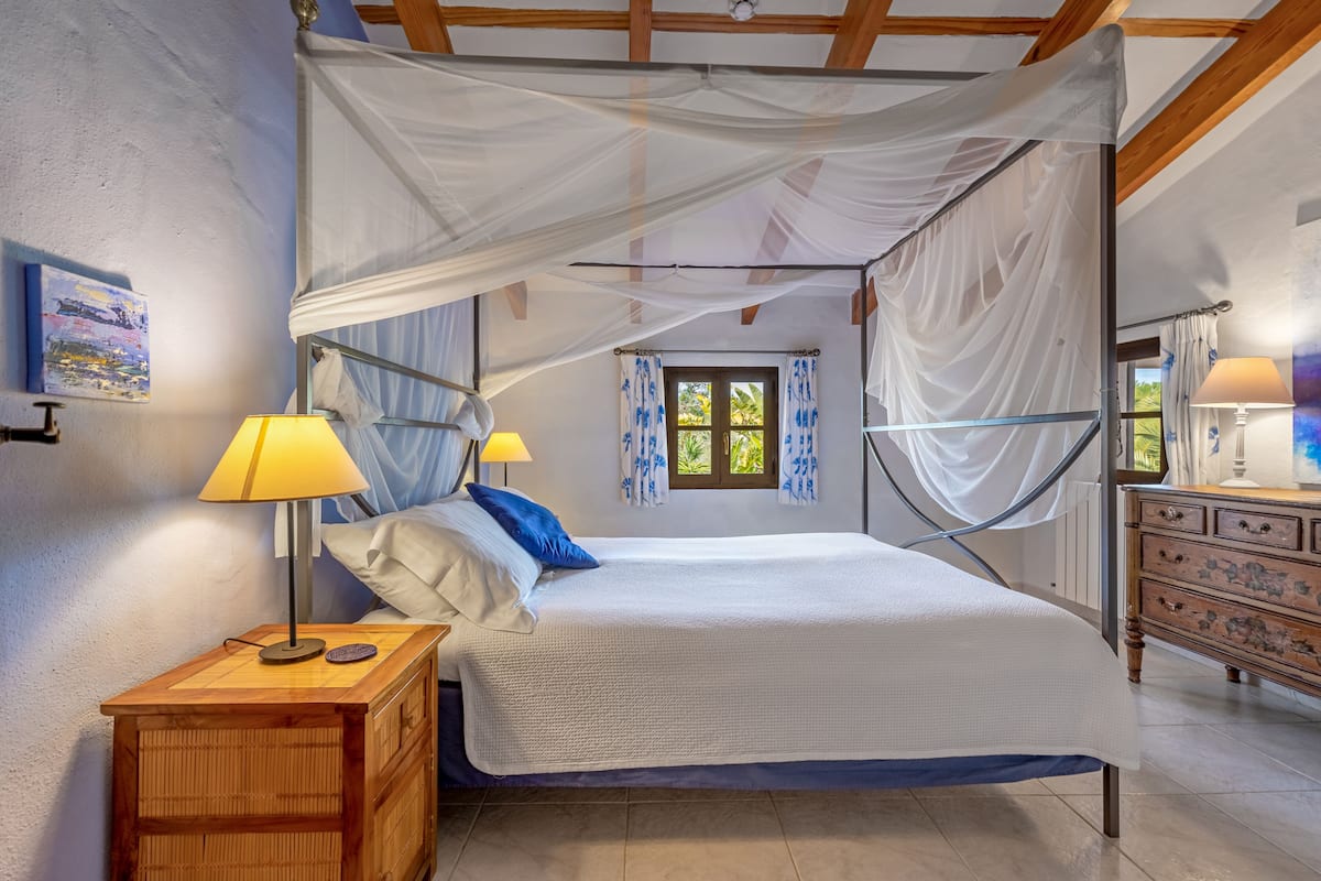 Quarto com cama de dossel, mesas de cabeceira de madeira, cômoda e janela com cortinas.