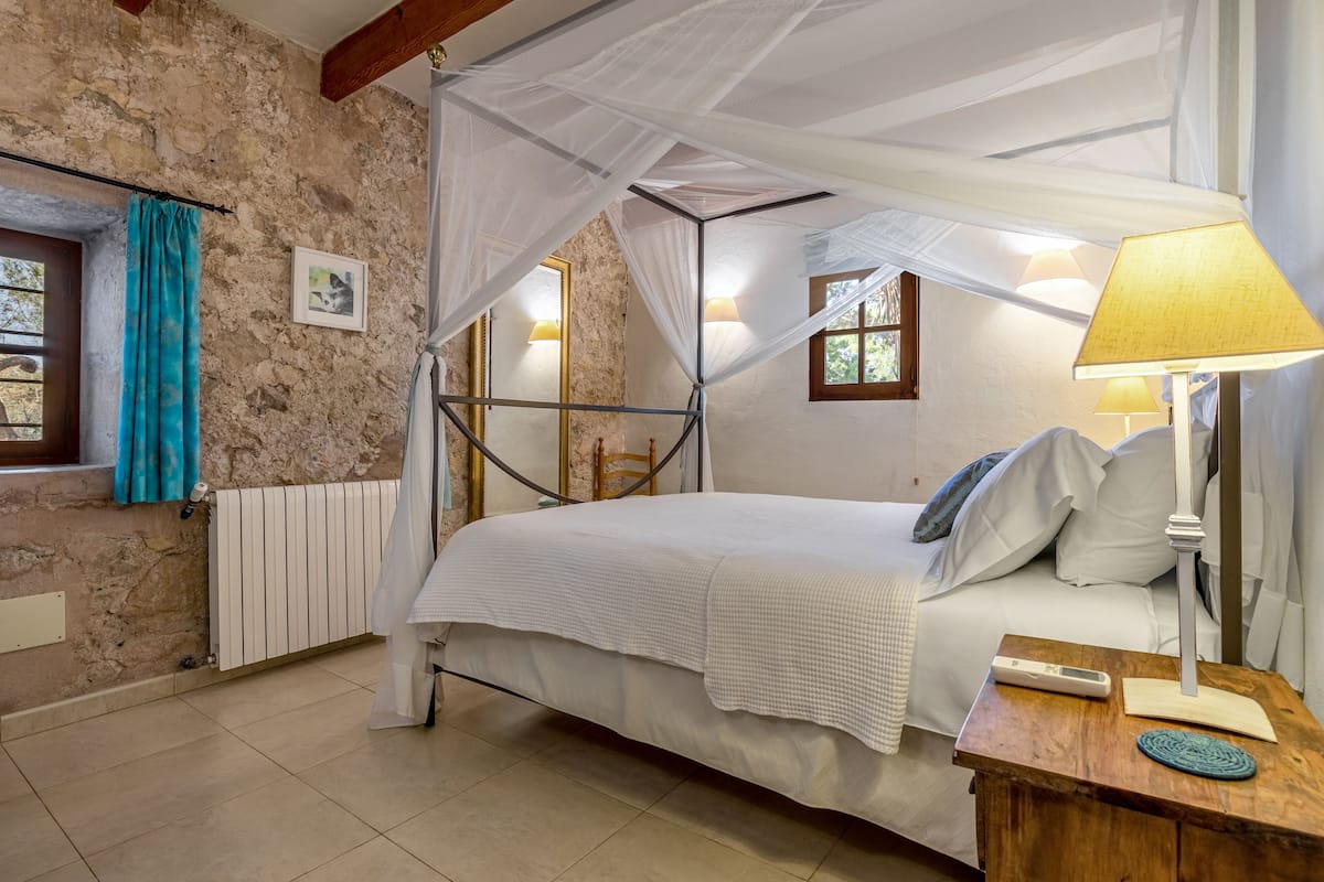 Quarto com cama de dossel, luminária de cabeceira, mesa de cabeceira de madeira e janela com cortinas.