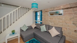 A modern living room with a grey sectional sofa, a small white side table, a blue rug, a staircase with white railings, and a stone wall. - Kirkstall Bridge Apartments - 26 (Leeds)