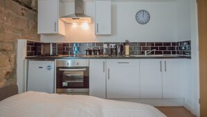 A compact kitchen with white cabinets, a built-in oven, a microwave, and a clock on the wall. - Kirkstall Bridge Apartments - 26 (Leeds)