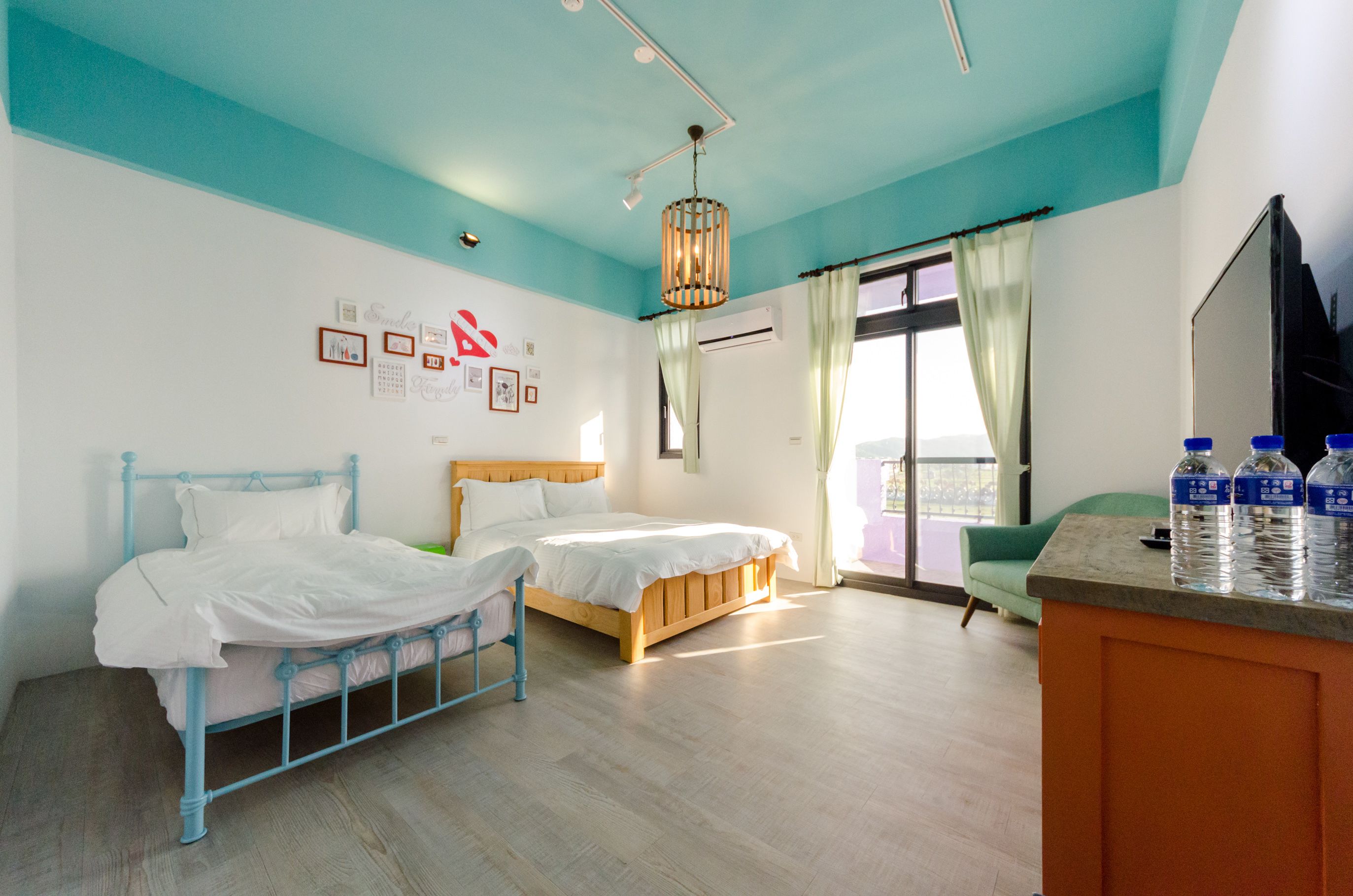 A hotel room with two beds, a wooden headboard, a large window with curtains, a green chair, and a minibar with bottled water.