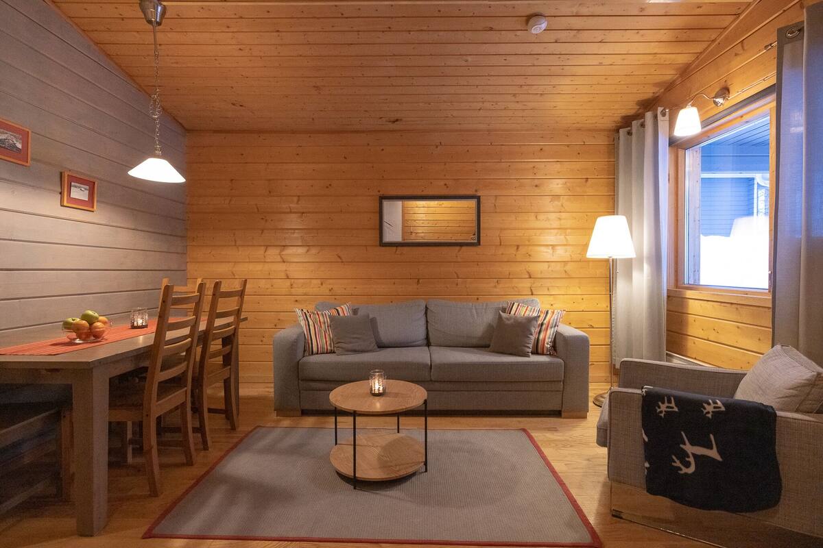 A cozy room with a wooden ceiling, a grey sofa, a round coffee table, and a dining area with chairs and a table.