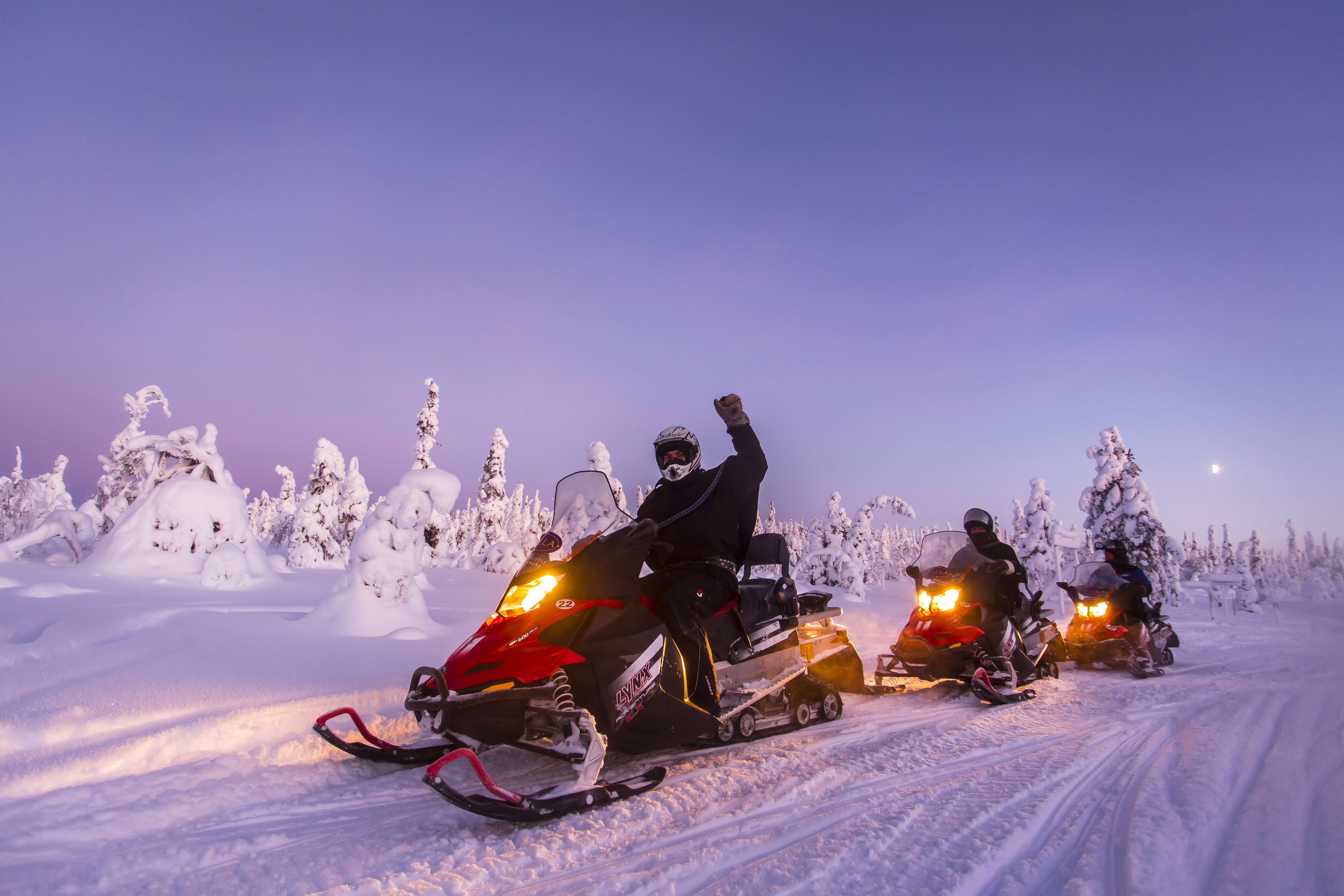 Três motos de neve em um caminho coberto de neve, com árvores carregadas de neve e céu claro.