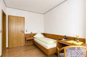 A small, single-bed room with a wooden door, a desk with a chair, a bedside table, and a small potted plant. - Landhotel Hamburger Hof (Söhlde)