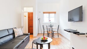 Un salon moderne avec un canapé en cuir noir, une table ronde en bois sur laquelle est posé un saladier de fruits, un coin repas avec une table et des chaises en bois, et un téléviseur fixé au mur.
