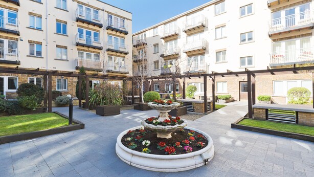 Une cour intérieure avec une fontaine centrale, entourée de parterres de fleurs, d’une pergola et d’immeubles résidentiels à plusieurs étages dotés de balcons.
