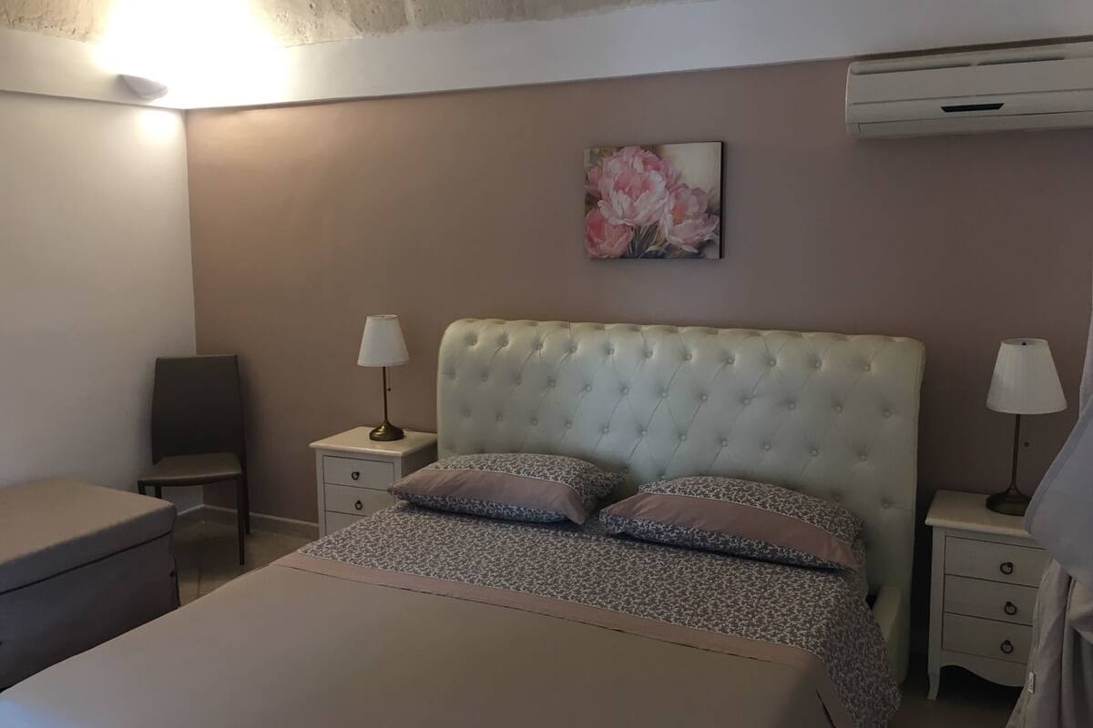 A bedroom with a tufted headboard, two bedside tables with lamps, a floral artwork on the wall, and an air conditioning unit.