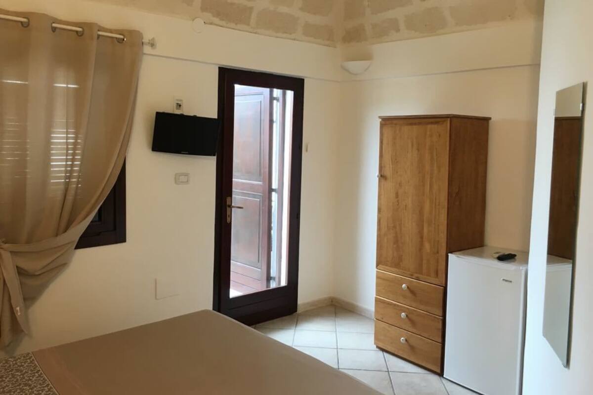 A room with a bed, a wooden wardrobe, a television mounted on the wall, and a door leading outside.